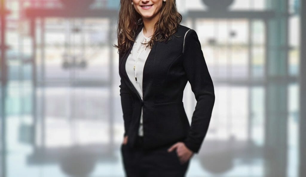 Une personne aux cheveux longs et bruns se tient debout, les mains dans les poches, souriant. Elle porte un blazer noir et un chemisier blanc. L'arrière-plan est flou, avec de grandes fenêtres et un intérieur moderne.