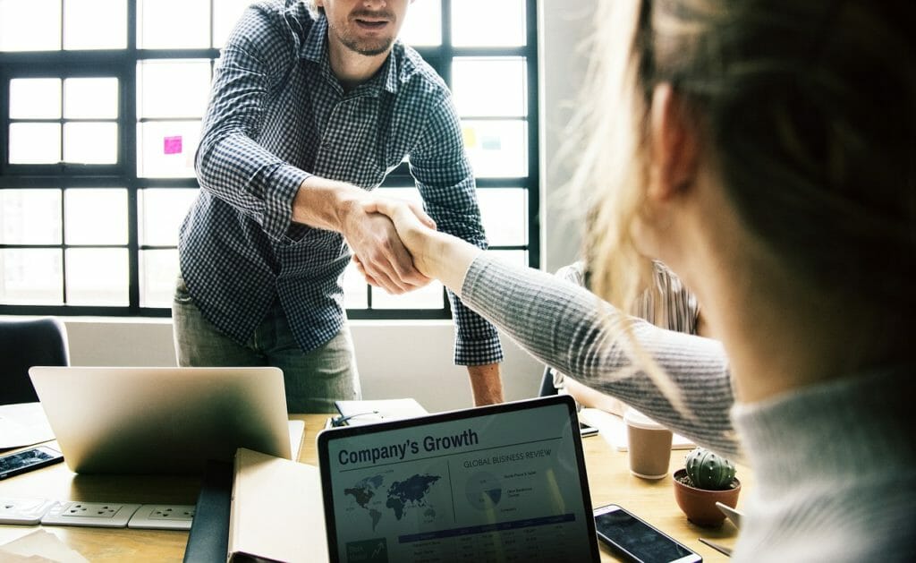 Un homme et une femme se serrent la main dans un bureau, signe d'un accord commercial ou d'une collaboration. Des ordinateurs portables, des papiers et une tasse sont posés sur une table en bois, avec une fenêtre en arrière-plan laissant entrer la lumière naturelle. L'écran d'un ordinateur portable affiche le graphique de croissance d'une entreprise.