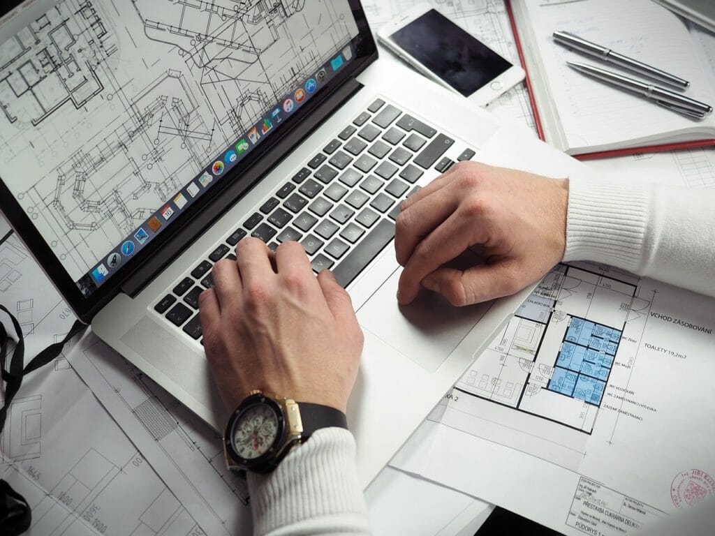 Une personne travaille sur un ordinateur portable affichant des plans d'architecture. Le bureau est encombré de plans imprimés supplémentaires, d'un smartphone, d'un carnet et d'un stylo. La personne porte une montre et une chemise blanche à manches longues.