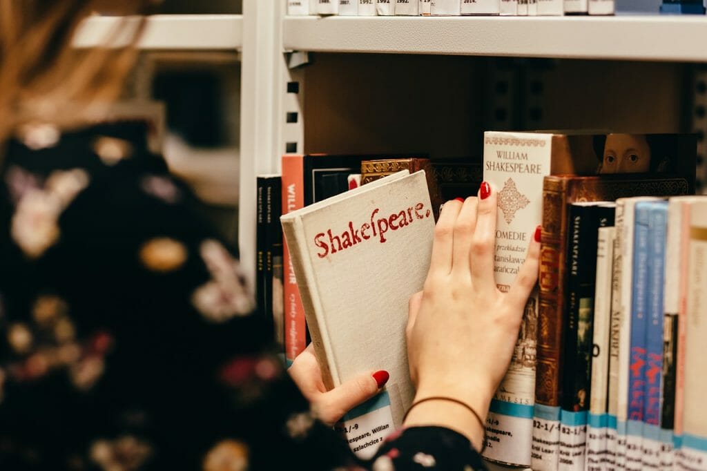 Une personne au vernis à ongles rouge sort un livre blanc intitulé « Shakespeare » d’une étagère remplie de livres divers, dont « Qu’est-ce que la Pédagogie Inversée ? ». Seules sa main et une partie de son bras sont visibles, encadrées par d’autres livres et l’étagère en arrière-plan.