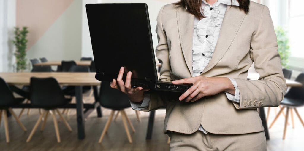 Une personne en costume beige tenant un ordinateur portable ouvert se tient dans un bureau avec des chaises et des tables floues en arrière-plan. L'environnement de bureau semble moderne avec des sols en bois et des murs de couleur claire. Seule la moitié supérieure du corps de la personne est visible.