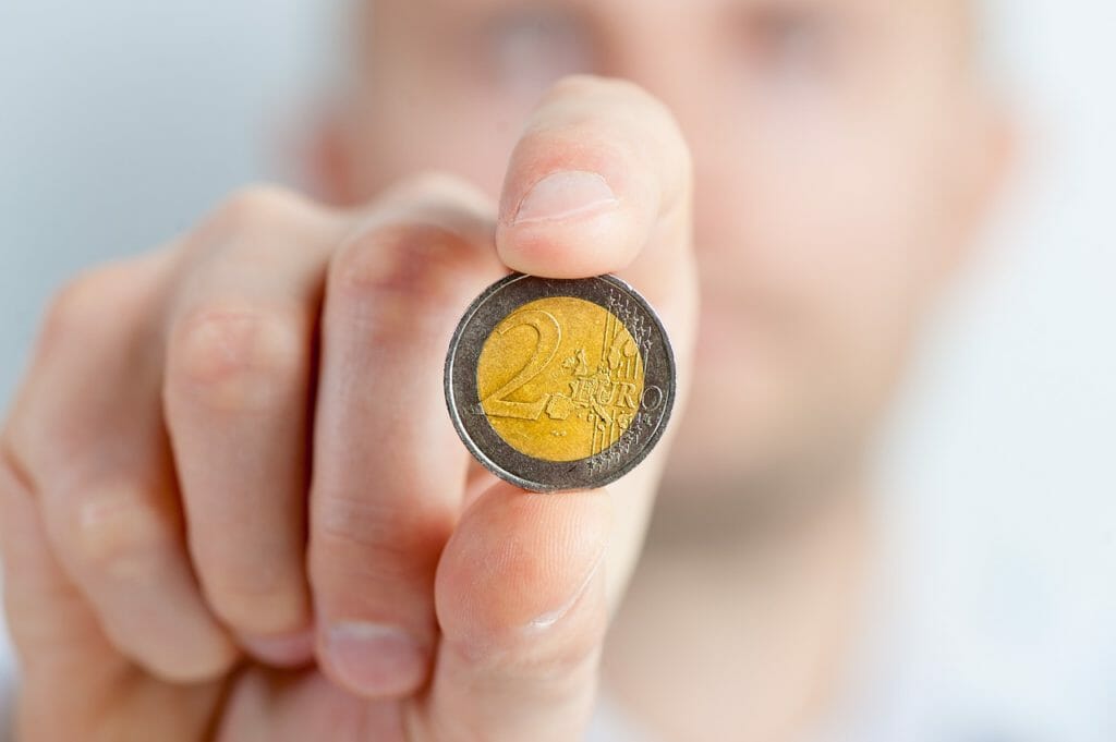 Une personne tient une pièce de 2 euros entre le pouce et l'index, la pièce étant tournée vers l'objectif et la personne étant floue en arrière-plan. Le dessin et la valeur de la pièce sont clairement visibles.