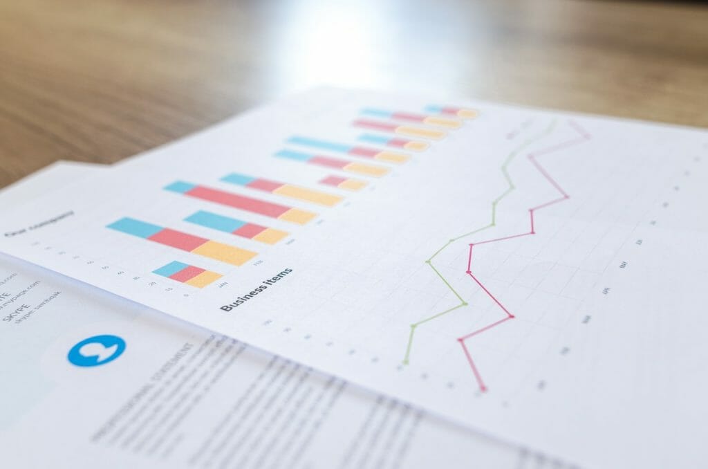 Gros plan de documents commerciaux imprimés sur une surface en bois, avec des graphiques à barres et à lignes colorés. Le graphique à barres affiche des données segmentées en bleu, orange et rouge, tandis que le graphique à lignes suit les tendances avec des lignes rouges et vertes. Le texte est partiellement visible sur les pages.