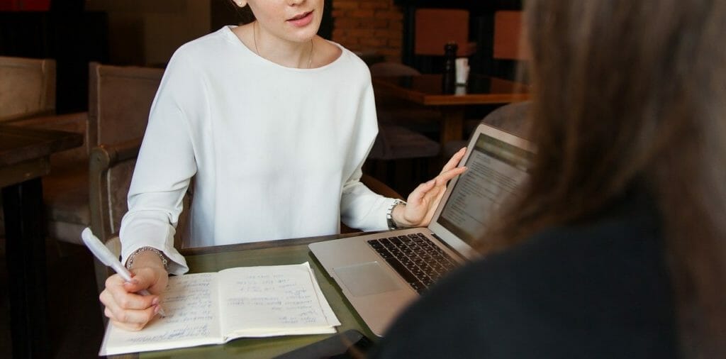 Deux femmes sont assises à une table et discutent. L'une d'elles, au premier plan, tient un stylo et consulte un carnet comme si elle prenait des notes. L'autre femme, vue de dos, semble écouter attentivement tout en faisant face à un ordinateur portable.