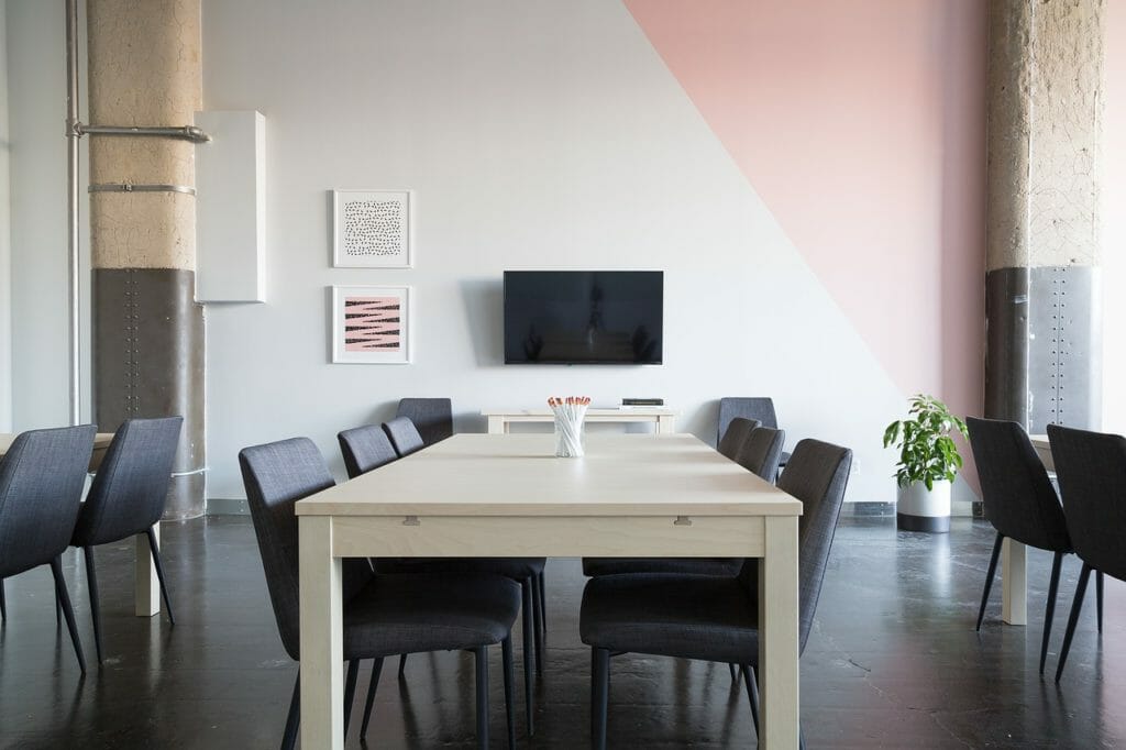 Une salle de conférence moderne avec une table beige et des chaises noires. Un grand téléviseur à écran plat est fixé sur un mur gris clair aux accents géométriques roses. Des œuvres d'art abstraites et une petite plante ornent le mur de manière décorative. Une grande plante verte est placée près du mur de droite.