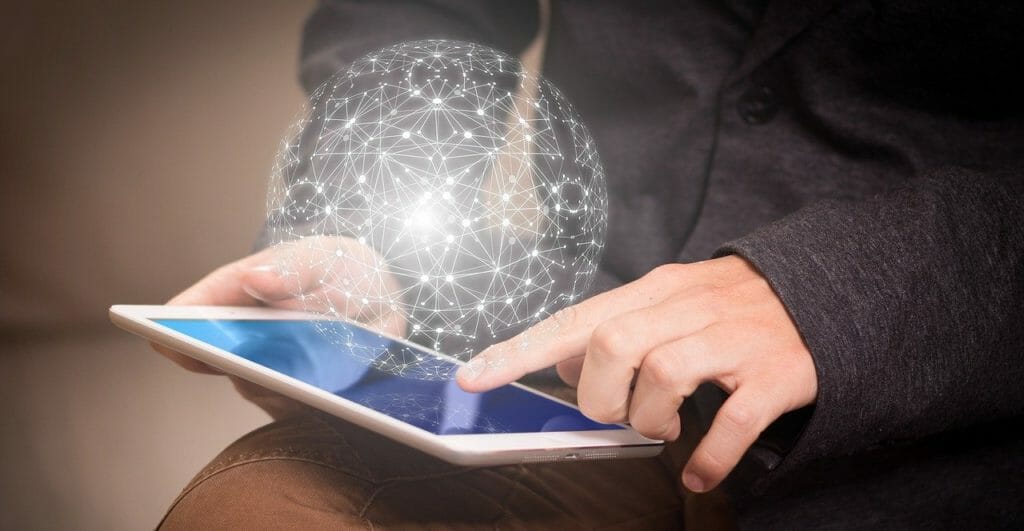 Une personne utilise une tablette d'une main tout en touchant l'écran de l'autre. Un globe holographique semi-transparent et lumineux flotte juste au-dessus de l'écran de la tablette. La tenue de la personne suggère un cadre formel ou professionnel.