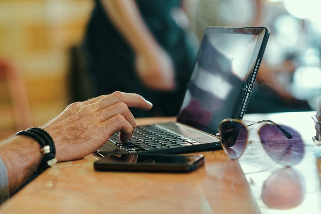 Une personne tape sur un ordinateur portable à une table, avec un smartphone et des lunettes de soleil posés à côté d'elle. L'arrière-plan est flou, concentrant la scène sur l'ordinateur portable et d'autres objets sur la table.