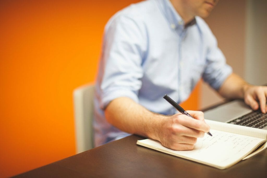Une personne vêtue d'une chemise bleu clair est assise à un bureau, écrivant dans un cahier avec un stylo. Un ordinateur portable est ouvert devant elle et l'arrière-plan présente un mur orange. Le décor semble être celui d'un bureau ou d'un espace de travail décontracté.