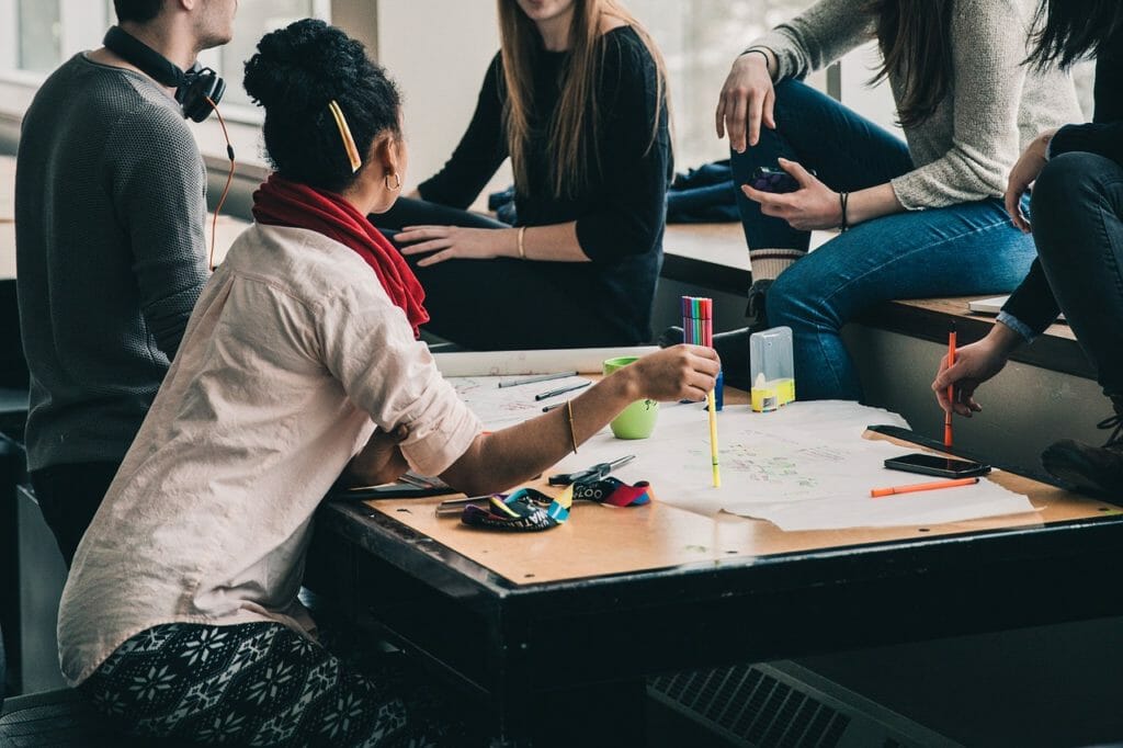 Un groupe de personnes est réuni autour d'une table et discute. La table est encombrée de papiers, de crayons et de marqueurs colorés. Une personne écrit, tandis que d'autres sont assises ou penchées à côté. La scène suggère une séance de collaboration ou de brainstorming.