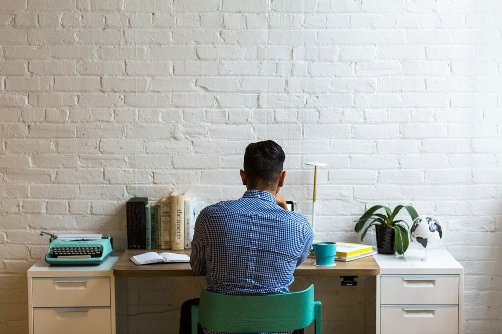 Une personne vêtue d'une chemise à carreaux bleus est assise à un bureau face à un mur de briques blanches. Le bureau est équipé d'une machine à écrire, de livres, d'un cahier, d'une lampe, d'une tasse, d'une plante en pot et d'un petit globe.