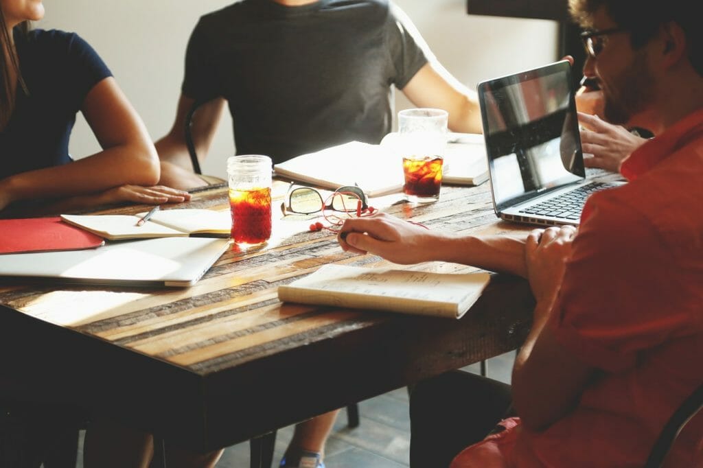 Un groupe de personnes assises autour d'une table en bois, en pleine discussion. Des ordinateurs portables, des cahiers et des verres de boissons glacées sont posés sur la table. Le cadre semble être celui d'une réunion informelle ou d'une séance de brainstorming dans une pièce bien éclairée.