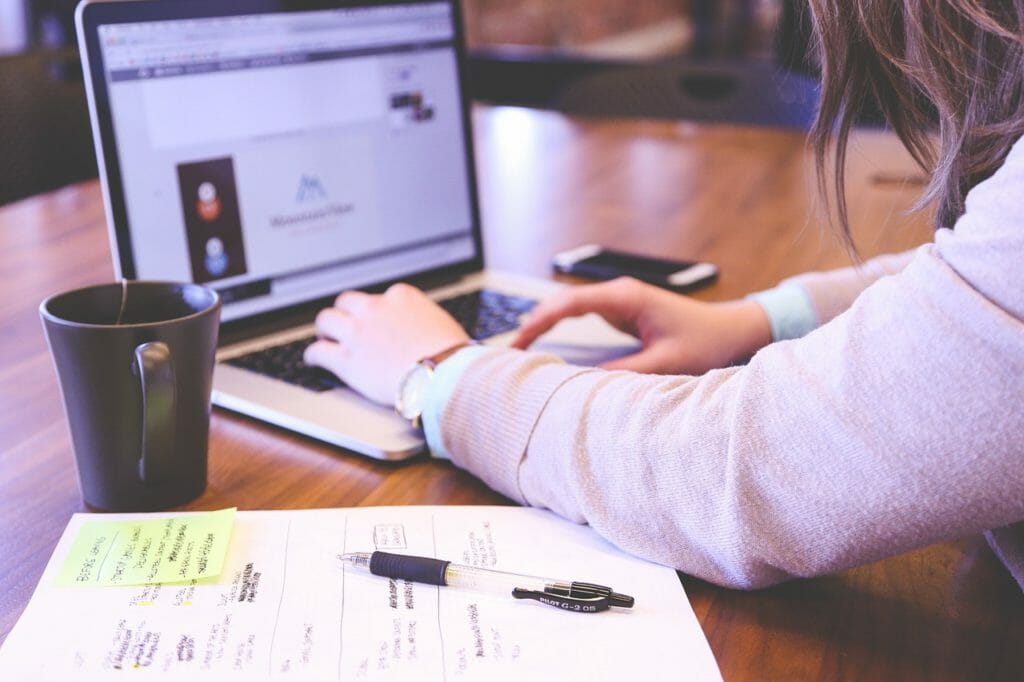 Une personne tape sur un ordinateur portable posé sur un bureau en bois. À côté de l'ordinateur portable, il y a une tasse à café noire. Un bloc-notes avec des notes manuscrites, un stylo et un smartphone sont également posés sur le bureau. L'écran affiche une page Web avec des graphiques et du texte.