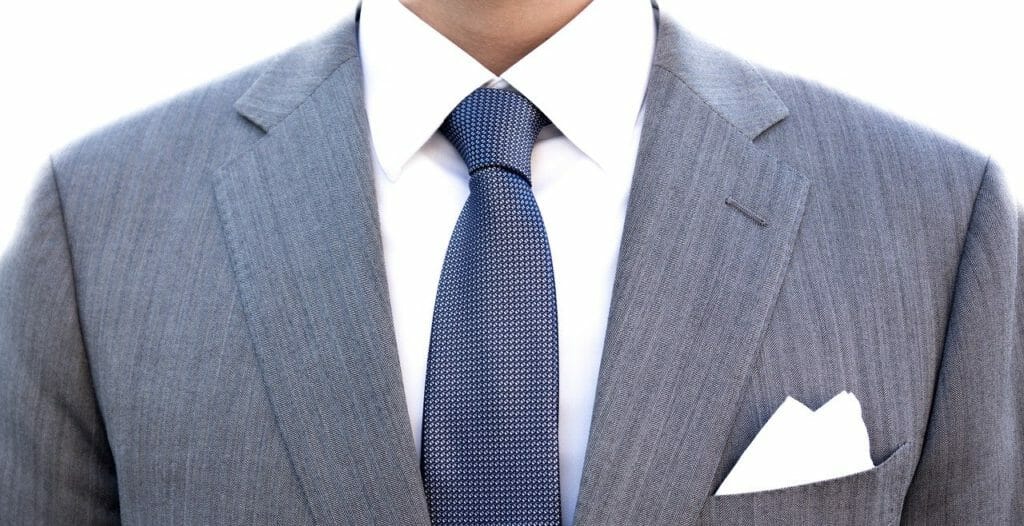 Une personne porte une veste de costume gris clair avec une chemise blanche et une cravate bleue. Une pochette blanche est soigneusement glissée dans la poche de poitrine de la veste. L'arrière-plan est blanc uni. Le visage de la personne n'est pas visible sur l'image.