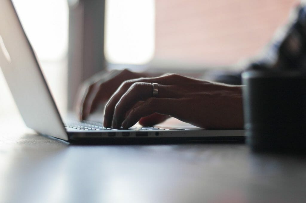 Gros plan d'une personne tapant sur un clavier d'ordinateur portable. La personne porte une bague à l'annulaire et l'arrière-plan est légèrement flou, ce qui permet de mettre en valeur les mains et le clavier. L'éclairage est doux, créant une atmosphère calme.