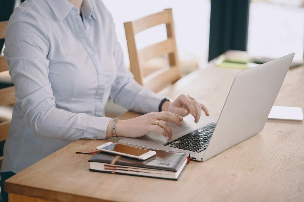 Une personne en chemise blanche tape sur un ordinateur portable assis à une table en bois. Un smartphone, un cahier et un stylo sont posés sur la table à côté de l'ordinateur portable. L'arrière-plan comprend une autre chaise et une fenêtre avec de la lumière naturelle.