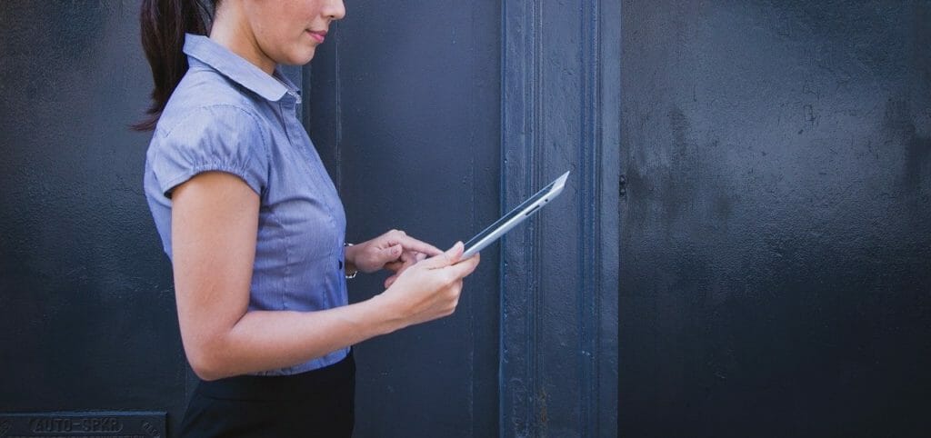 Une personne portant une chemise bleu clair à manches courtes se tient debout contre un mur sombre, tenant et utilisant une tablette. Seul le haut du corps de la personne est visible de profil. Il y a un léger reflet sur l'écran de la tablette.
