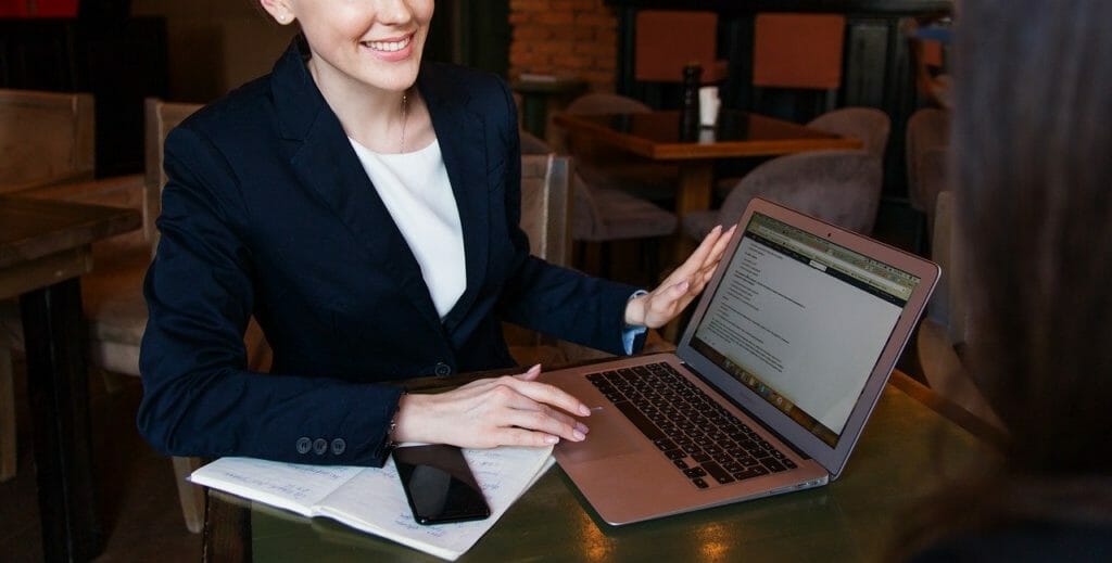 Une personne en tenue professionnelle est assise à une table dans un café, souriante et regardant l'écran d'un ordinateur portable. Un smartphone et un bloc-notes avec un stylo sont posés sur la table à côté d'elle. Le café a une ambiance détendue avec des meubles en bois et un éclairage tamisé.