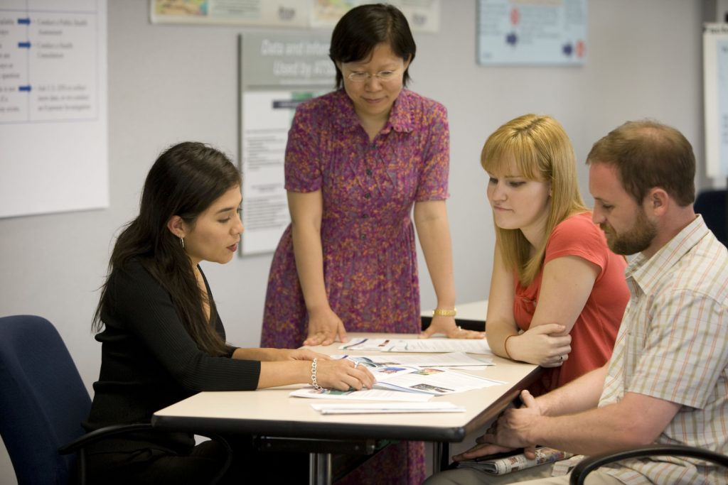 Un groupe de quatre personnes, composé de deux femmes et de deux hommes, est réuni autour d'une table pour discuter de documents. Une femme se lève et observe tandis que les autres sont assises et examinent les documents. Le décor semble être celui d'un bureau ou d'une salle de réunion avec des affiches sur les murs.
