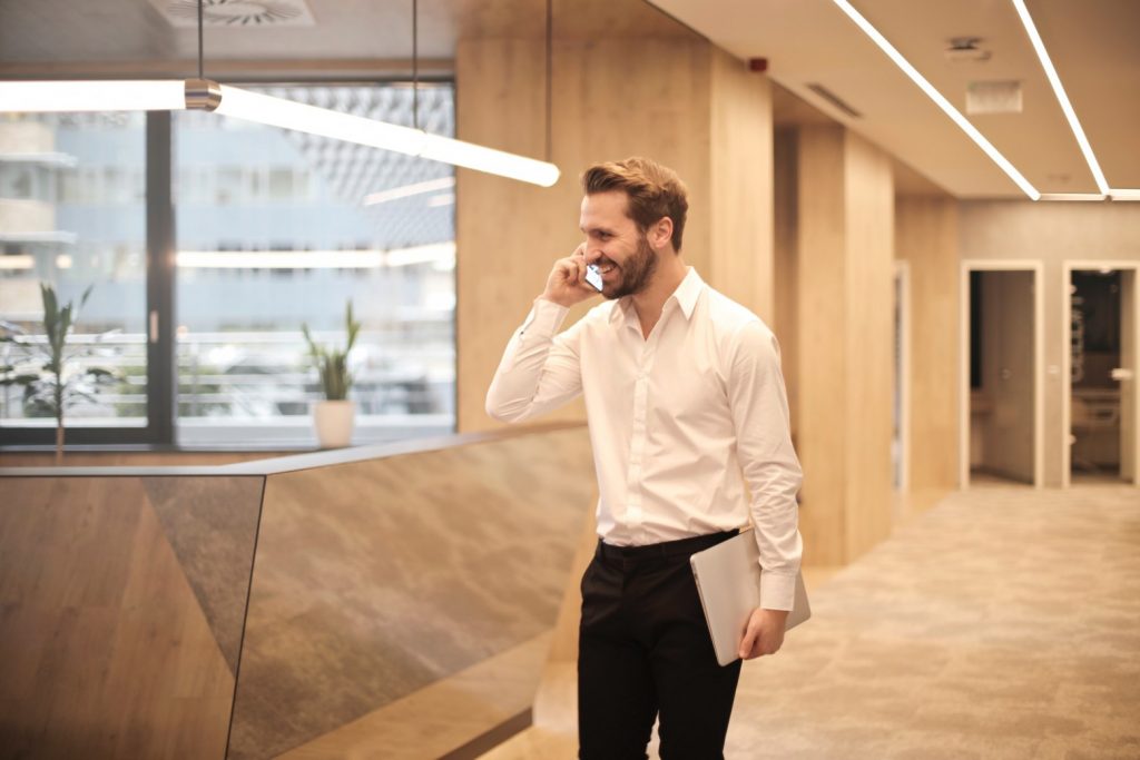 Un homme portant une chemise blanche et un pantalon noir sourit tout en parlant au téléphone. Il tient un ordinateur portable dans son autre main. L'arrière-plan, montrant un bureau moderne avec de grandes fenêtres et une décoration en bois, suggère qu'il pourrait discuter de sujets tels que « Qui finance le CPF 2020 ?