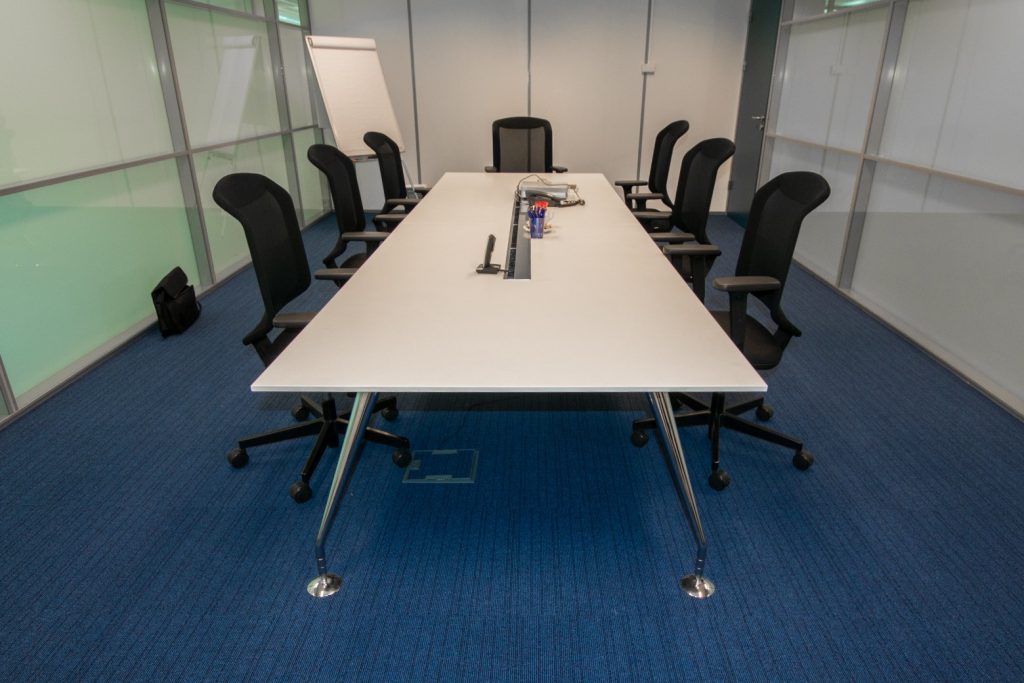 Une salle de conférence moderne avec une longue table blanche entourée de sept chaises de bureau noires sur un sol en moquette bleue. La table est équipée de prises de courant intégrées et abrite quelques fournitures de bureau. Un tableau à feuilles mobiles se trouve dans un coin et les murs environnants sont principalement en verre.