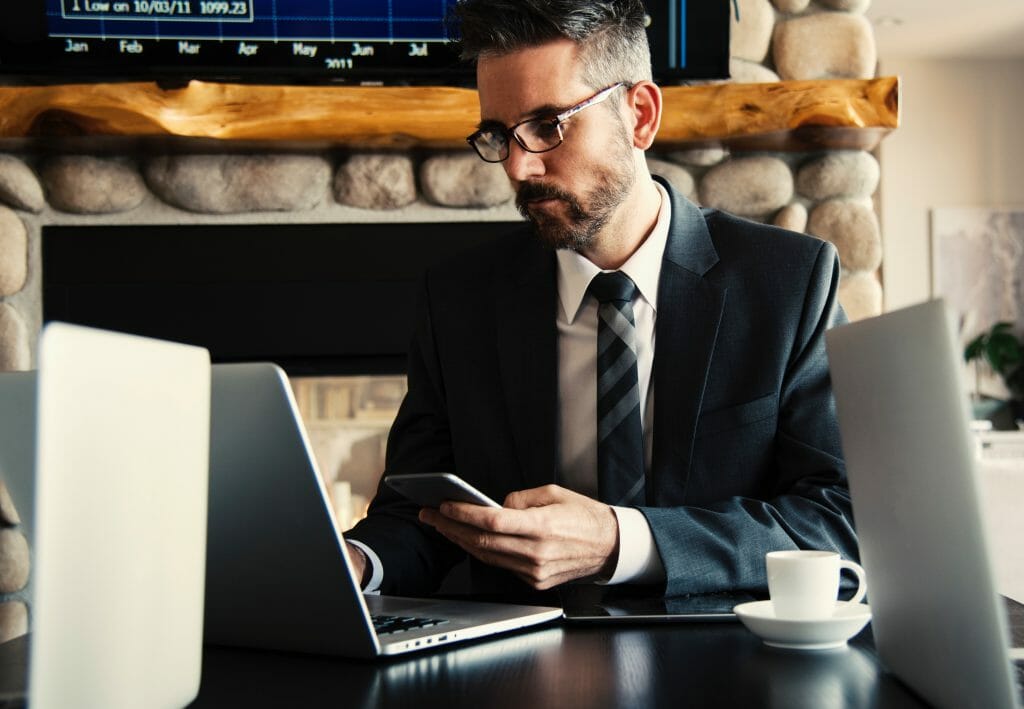 Un homme en costume-cravate est assis à un bureau, concentré sur son smartphone dans une main et tapant sur un ordinateur portable avec l'autre. Il est entouré d'un autre ordinateur portable, d'une tablette et d'une tasse de café. Une cheminée en pierre et un grand écran avec des graphiques sont visibles derrière lui, illustrant "Comment devenir Community manager sans diplôme ?".