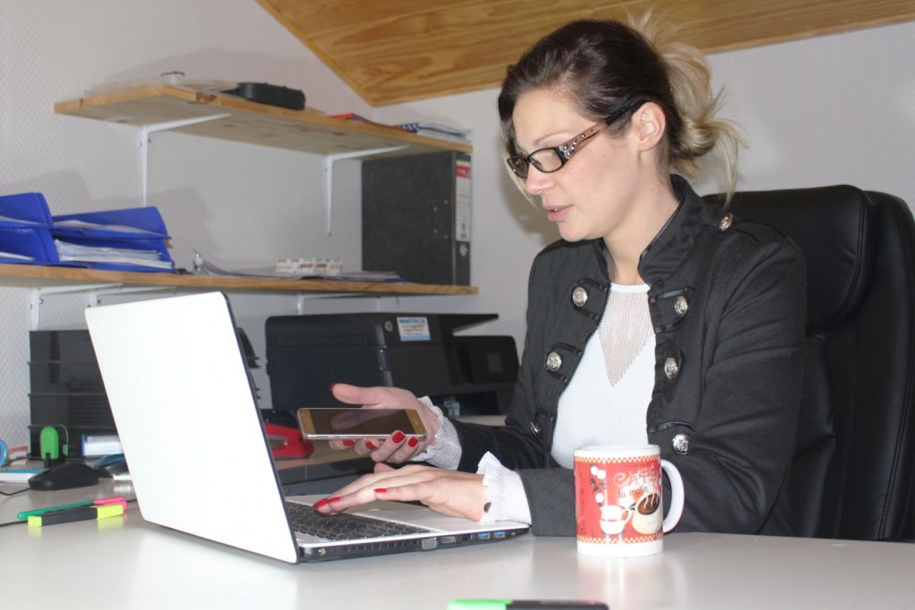 Une personne portant des lunettes et une veste noire est assise à un bureau, utilise un ordinateur portable et tient un smartphone, se demandant peut-être : « C’est quoi un Chef de projet numérique ? » Une tasse à café au motif festif est posée à côté de l’ordinateur portable, tandis que des étagères remplies de fournitures de bureau sont visibles en arrière-plan.
