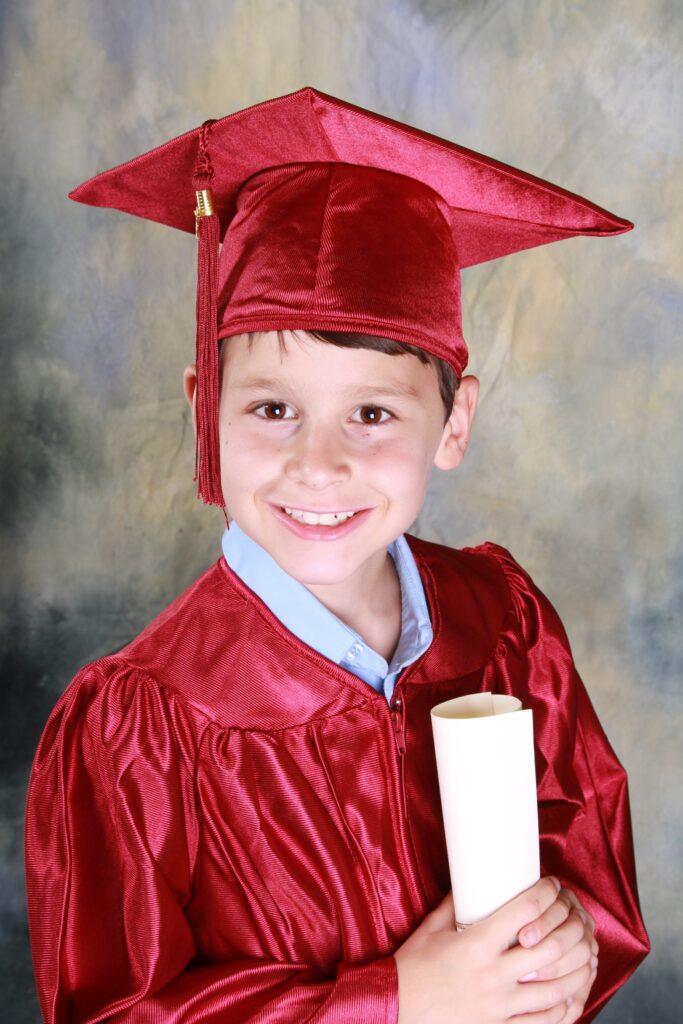 Un jeune garçon sourit tout en portant une toge et un chapeau de remise de diplôme rouges, tenant un diplôme roulé. L'arrière-plan présente un mélange de couleurs doux et abstrait.