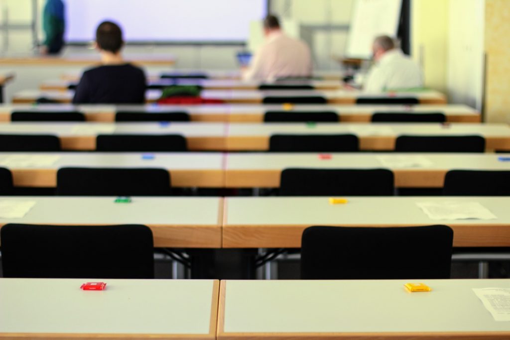 Une salle de classe avec des rangées de bureaux et de chaises vides, chaque bureau étant occupé par un objet d'une seule couleur. Trois personnes sont présentes à l'arrière-plan, dos à la caméra, deux assises à un bureau et une debout près d'un tableau blanc.