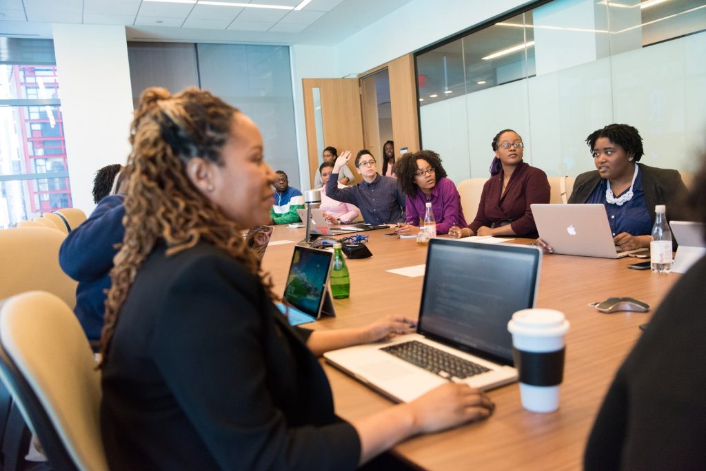 Un groupe de personnes discutent dans une salle de conférence moderne. Certains prennent des notes sur des ordinateurs portables, tandis que d'autres écoutent attentivement. La salle est dotée de grandes fenêtres et d'une longue table en bois avec divers objets personnels et boissons.