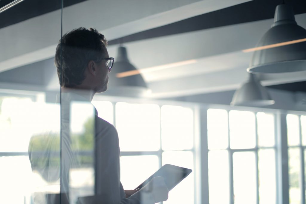 Une personne tenant une tablette se tient debout dans un bureau moderne doté de grandes fenêtres. La pièce est éclairée par la lumière naturelle et plusieurs luminaires fixés au plafond sont visibles. La personne semble être plongée dans ses pensées, le dos tourné vers la caméra.