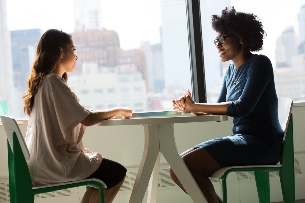 Deux femmes sont assises à une table près d'une grande fenêtre, en pleine conversation. L'une d'elles, aux cheveux longs et foncés, porte un haut clair et semble expliquer quelque chose à propos de la certification Qualiopi à l'autre femme aux cheveux bouclés et portant une robe bleue. L'arrière-plan montre un paysage urbain avec des bâtiments.
