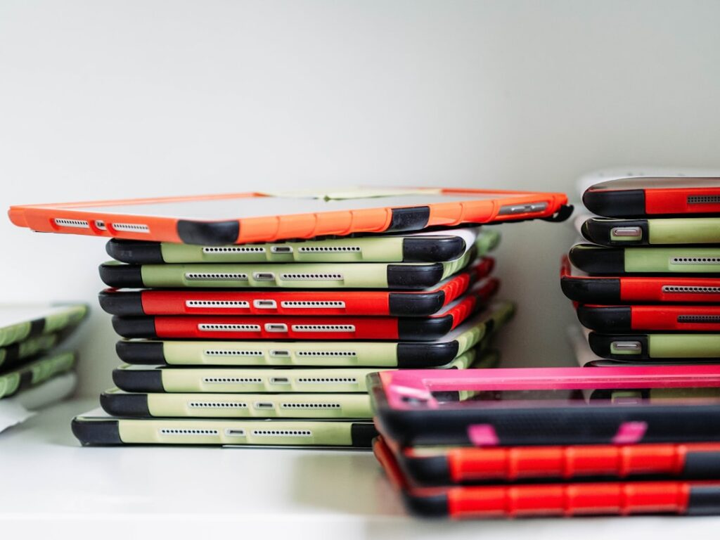 Une pile de tablettes dans des étuis de protection, principalement rouges et verts, est soigneusement disposée sur une étagère blanche. Les tablettes semblent être organisées en deux piles distinctes.