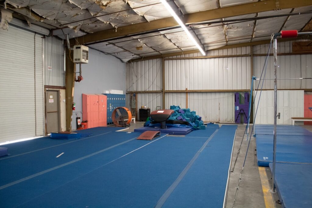 Un vaste espace d'entraînement de gymnastique avec un sol en tapis bleu, des barres parallèles asymétriques, une pile de blocs de mousse et des équipements de gymnastique contre les murs. Le plafond présente des signes d'usure et des casiers se trouvent sur la gauche.