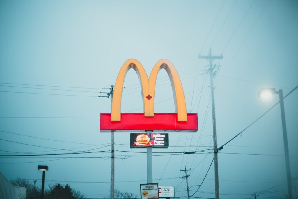 Le logo de McDonald's se dresse fièrement sur un ciel nuageux et brumeux. Les arches dorées sont bien visibles au sommet d'un panneau rouge. Des lignes électriques et des poteaux électriques sont visibles en arrière-plan, ce qui ajoute à l'ambiance industrielle.