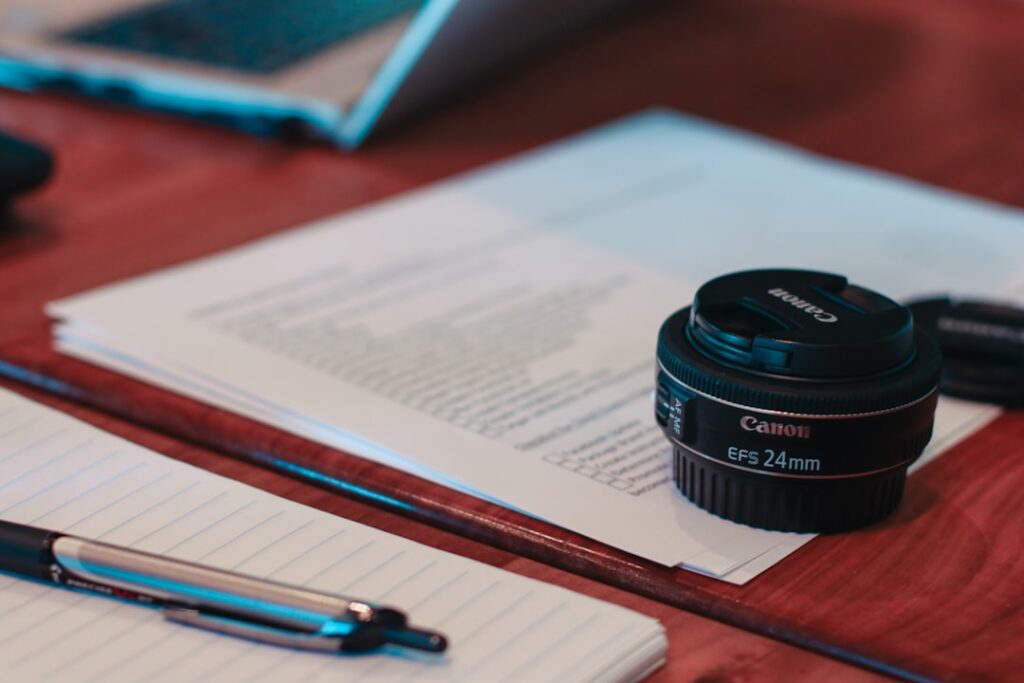 Un objectif Canon EFS 24 mm est posé sur une pile de documents imprimés sur une table en bois. À proximité, un stylo argenté est posé sur un cahier ligné. Une partie d'un ordinateur portable est visible en arrière-plan.