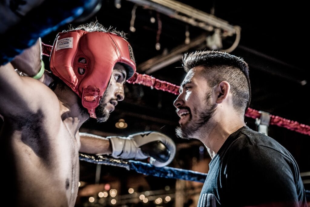 Un boxeur portant un casque rouge s'appuie contre les cordes d'un ring de boxe, tandis qu'un entraîneur lui parle avec une expression intense. Le décor est faiblement éclairé, ce qui contribue à l'atmosphère sérieuse.
