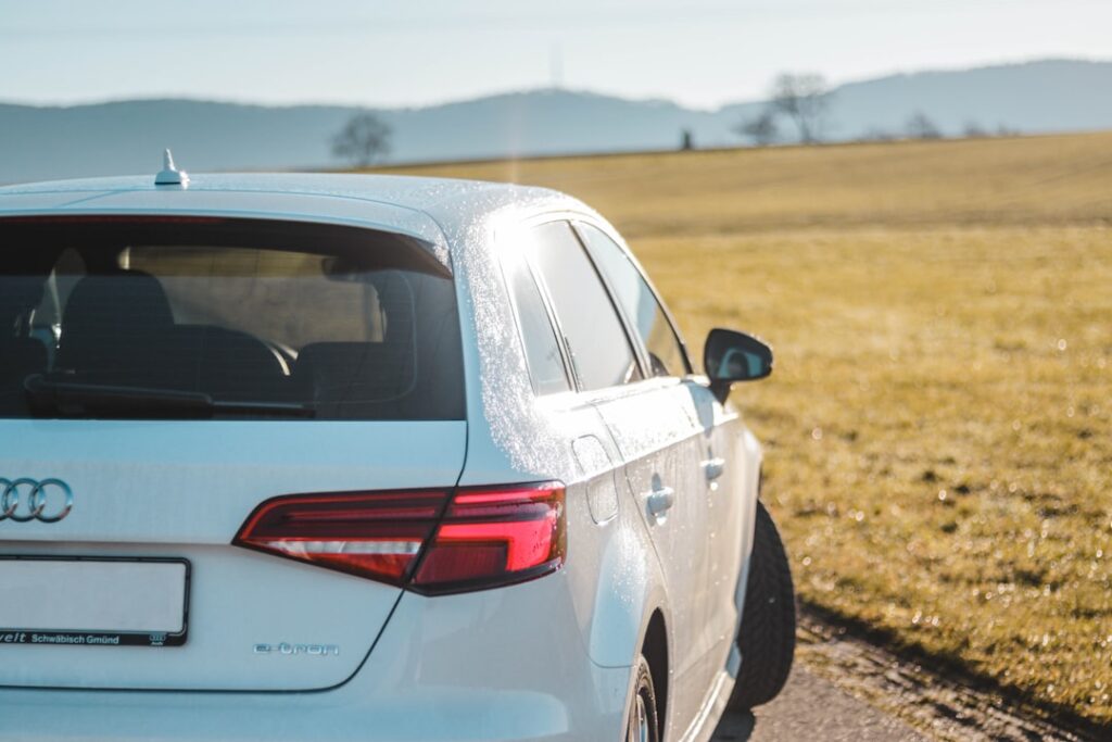 Une Audi e-tron blanche garée sur une route de campagne avec une vue panoramique sur les champs verdoyants et les collines lointaines sous un ciel bleu clair. La lumière du soleil se reflète sur la surface de la voiture, soulignant son design élégant.