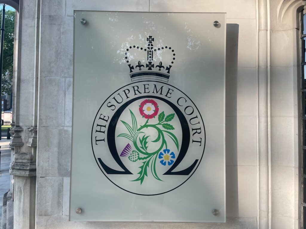 Panneau avec l'emblème de la Cour suprême, représentant un motif floral stylisé et une couronne, fixé sur un mur en pierre. L'emblème est circulaire, avec l'inscription « THE SUPREME COURT » tout autour.