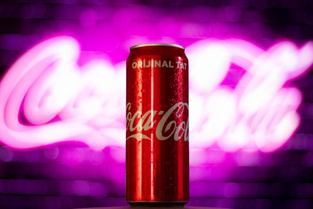 Une grande canette de Coca-Cola rouge avec de la condensation est centrée sur un fond rose et violet vibrant et flou comportant le logo Coca-Cola.