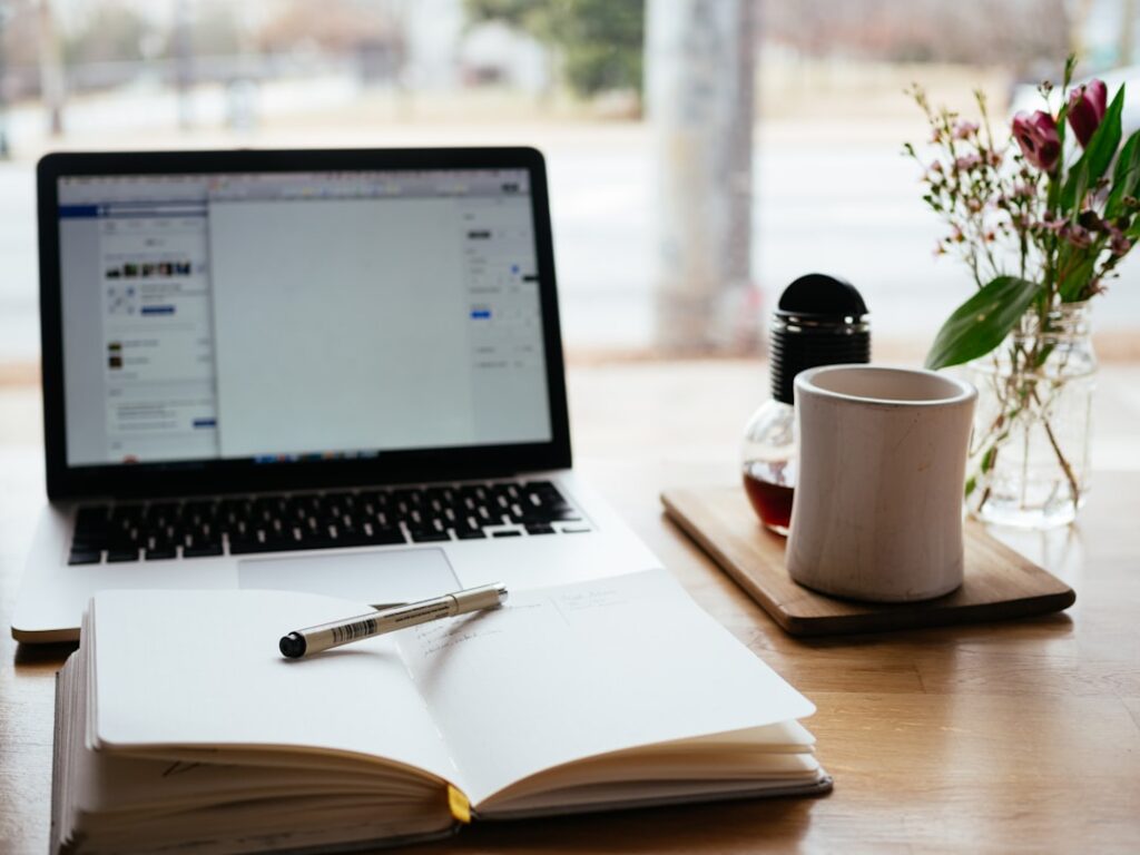 Un ordinateur portable ouvert sur une table en bois, affichant un document. Devant lui se trouve un cahier ouvert avec un stylo posé dessus. À droite, il y a une tasse blanche sur un plateau et un petit vase avec des fleurs. L'arrière-plan montre une vue extérieure floue.