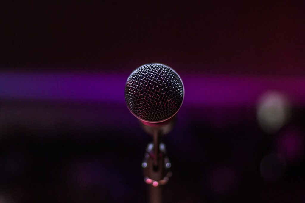 Gros plan d'un microphone sur un pied sur un fond flou et sombre avec un éclairage subtil violet et rouge, suggérant un décor de performance ou d'enregistrement.