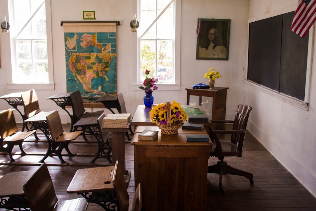 Une salle de classe vintage avec des bureaux en bois, un bureau de professeur décoré de fleurs, une carte des États-Unis au mur, un tableau noir et un drapeau américain. La lumière du soleil entre par les fenêtres, créant une atmosphère chaleureuse.