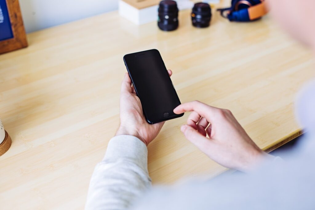 Une personne tient un smartphone dans une main et touche l'écran avec l'autre. L'écran du téléphone est éteint. La personne est assise à un bureau en bois clair avec des objectifs d'appareil photo en arrière-plan.