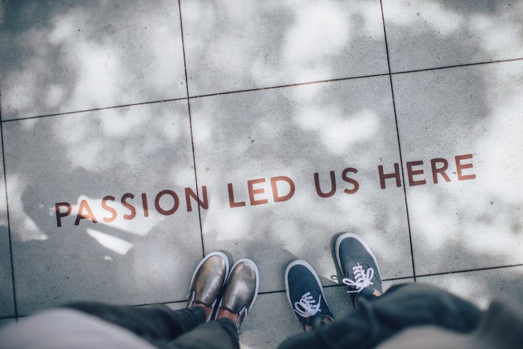 Deux personnes se tiennent sur un trottoir avec l'inscription « LA PASSION NOUS A CONDUIT ICI ». Leurs pieds, l'un chaussé de chaussures grises et l'autre de baskets bleues, sont visibles. Les ombres des arbres sont projetées sur le trottoir, créant un effet tacheté.