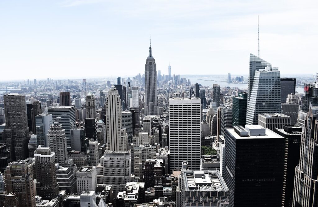 Vue panoramique de la ville de New York avec l'Empire State Building en plein centre, entouré d'autres gratte-ciels sous un ciel bleu clair. Le vaste paysage urbain de la ville s'étend jusqu'à l'horizon.