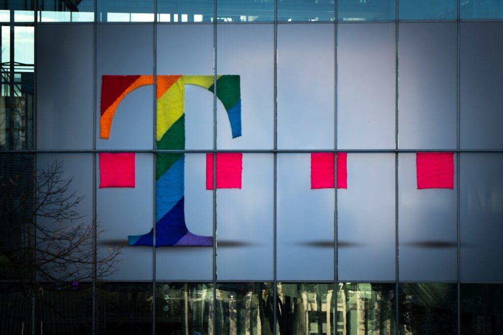 Un grand logo comportant la lettre « T » aux couleurs de l'arc-en-ciel est affiché sur la façade d'un bâtiment en verre, flanqué de deux carrés roses plus petits.