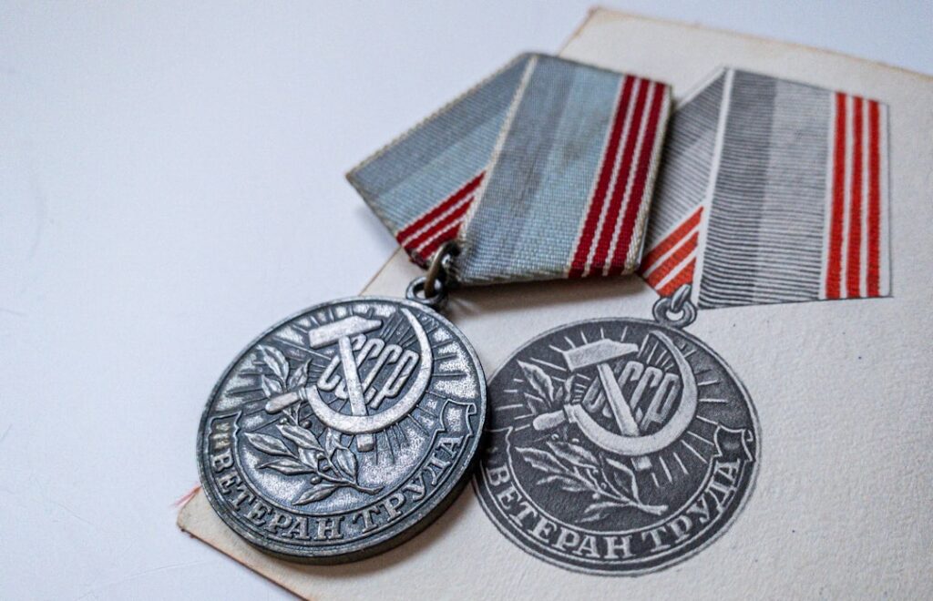 Deux médailles soviétiques anciennes avec des rubans rouges et blancs présentées sur une enveloppe. Chaque médaille présente un motif de faucille et de marteau avec un texte cyrillique. Les médailles reposent sur une surface légèrement inclinée, capturant un sentiment d'histoire et d'héritage.