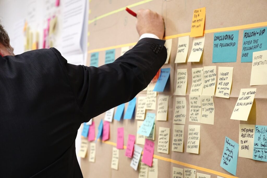 Une personne en costume écrit sur un grand tableau recouvert de notes autocollantes colorées contenant diverses notes et idées manuscrites. Le tableau semble être utilisé pour le brainstorming ou la planification de projets.
