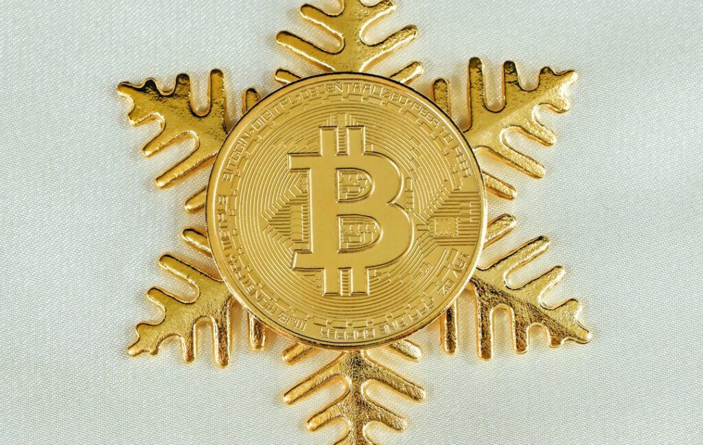 Une pièce Bitcoin en or positionnée au centre d'un flocon de neige décoratif en or, placé sur un fond de couleur claire.